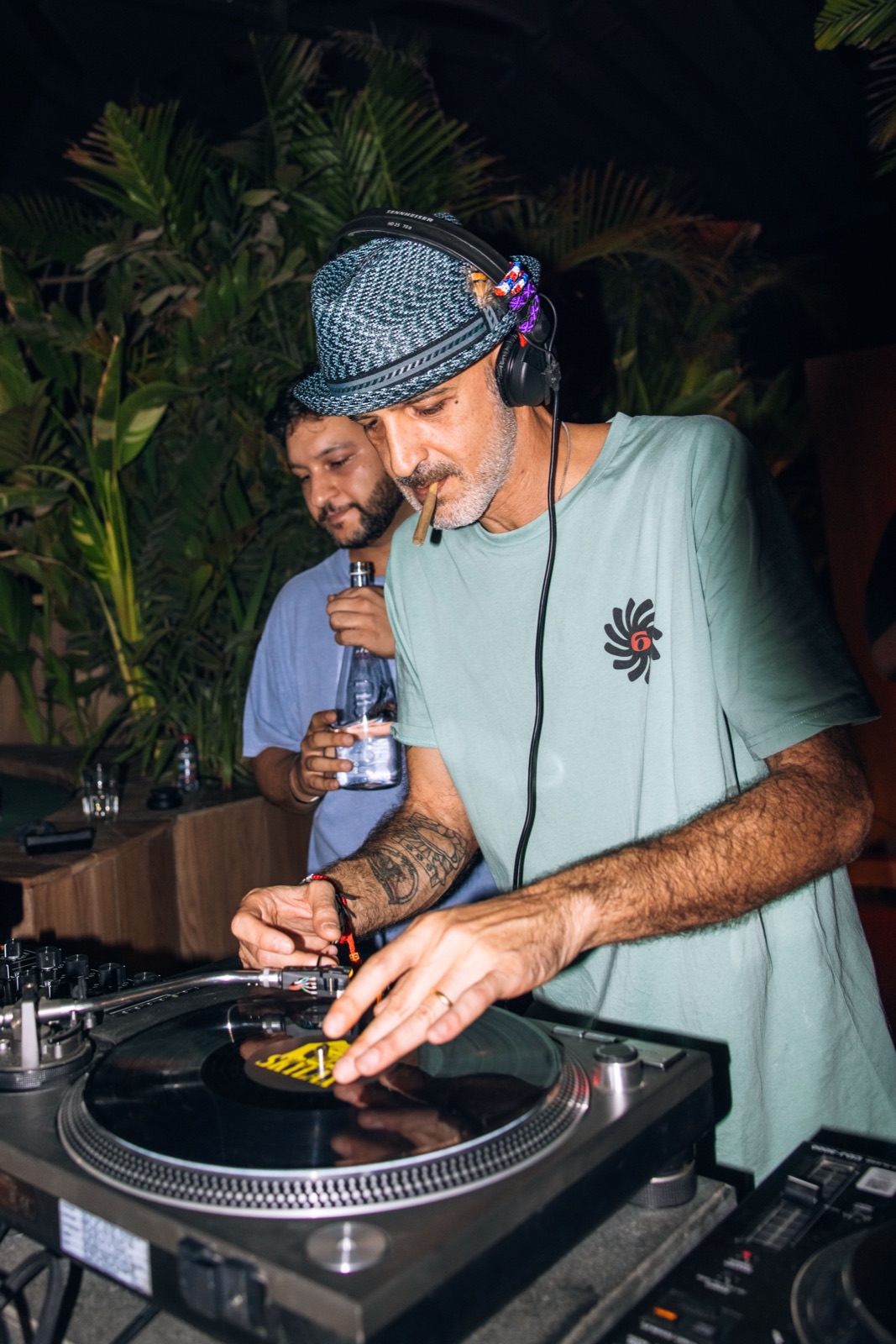 A DJ in a patterned hat cueing a record, with a second DJ behind.