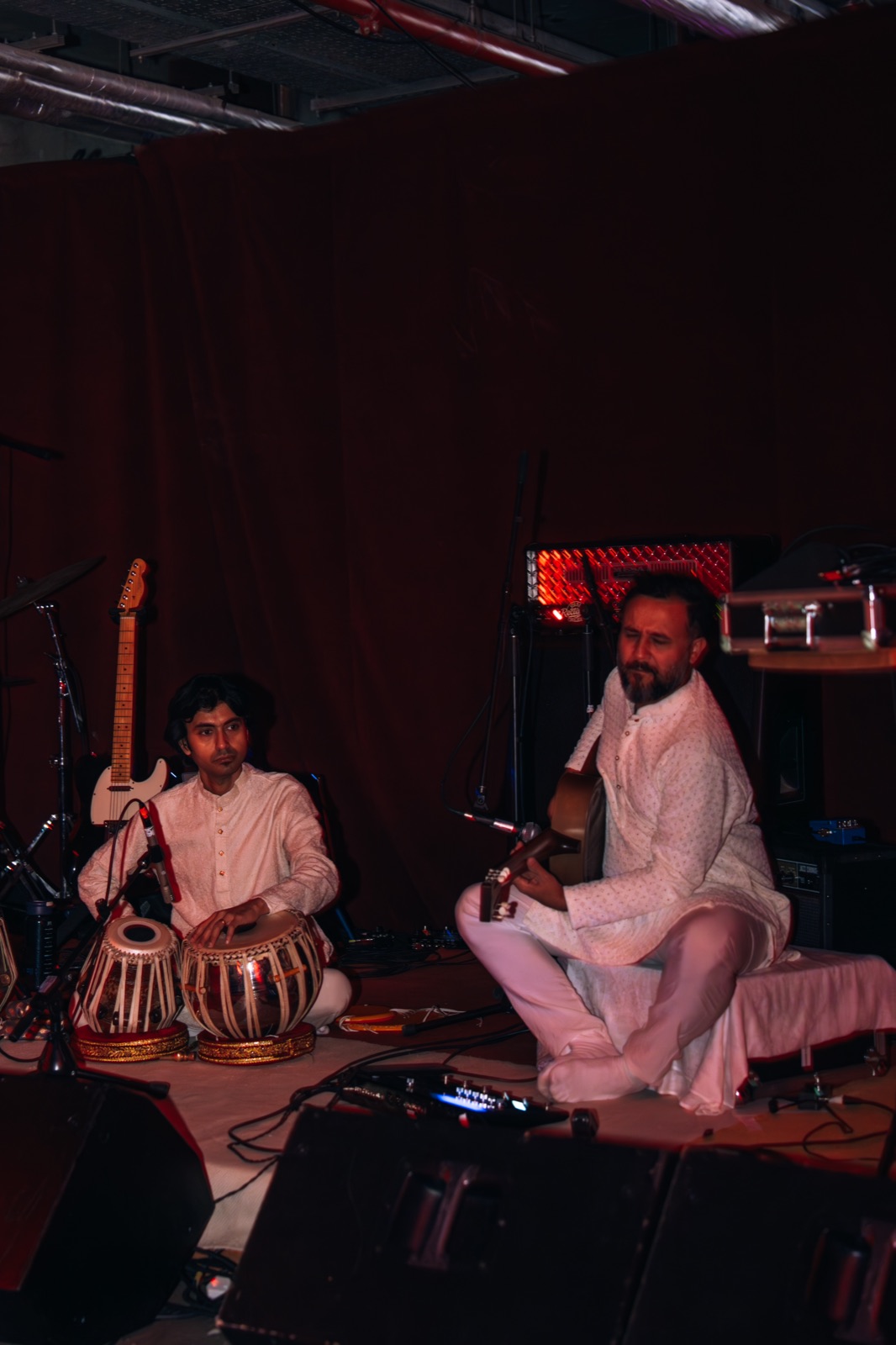 Two musicians on stage, one playing tabla and one a stringed instrument.
