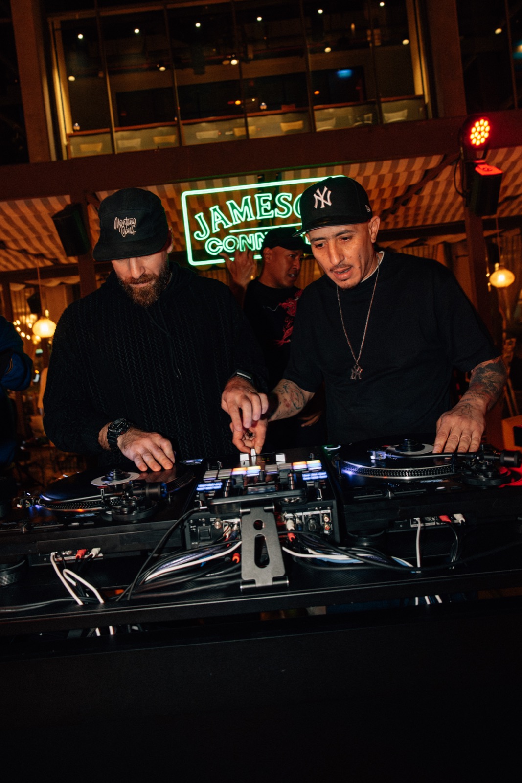 Two DJs side by side at the decks, one hand on the mixer.