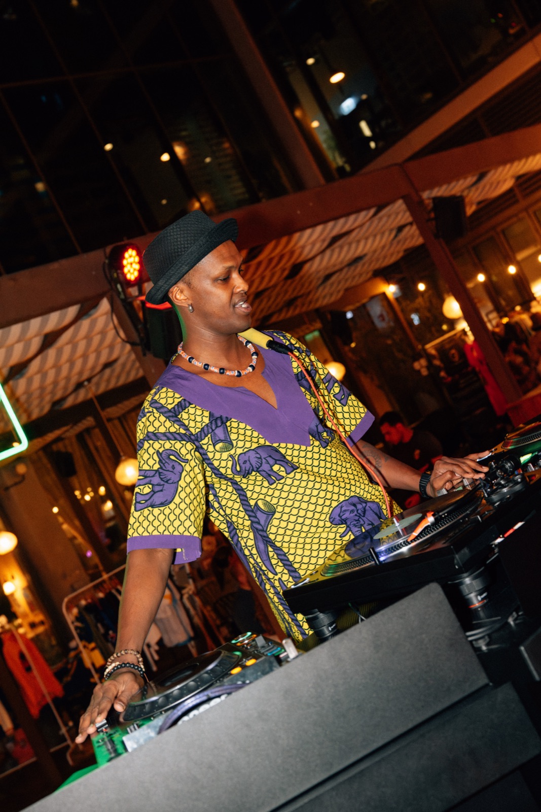 A DJ in a yellow and purple patterned shirt and a hat reaching across the decks.