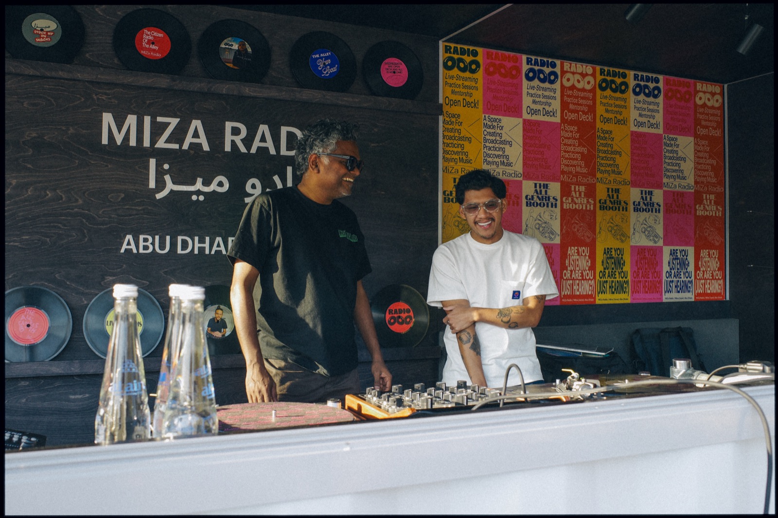 Two DJs at a booth in front of a Miza Radio Abu Dhabi sign and colourful posters.