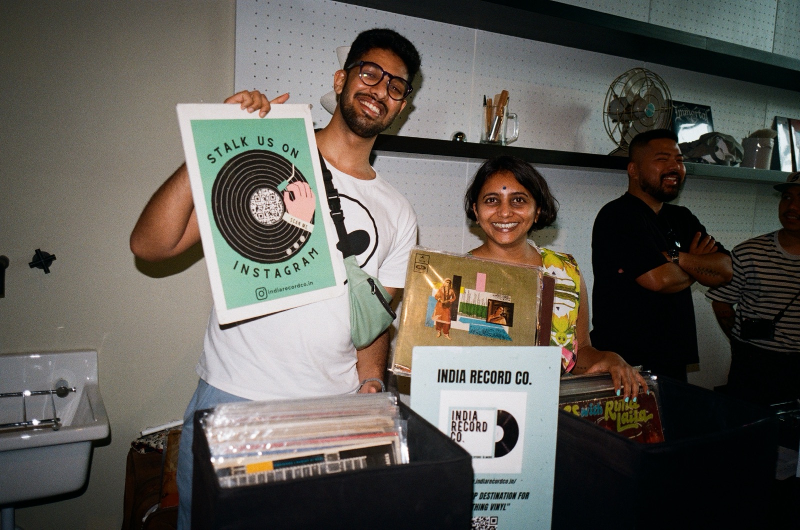 Two people smiling behind a stall, one holding up a record sleeve.