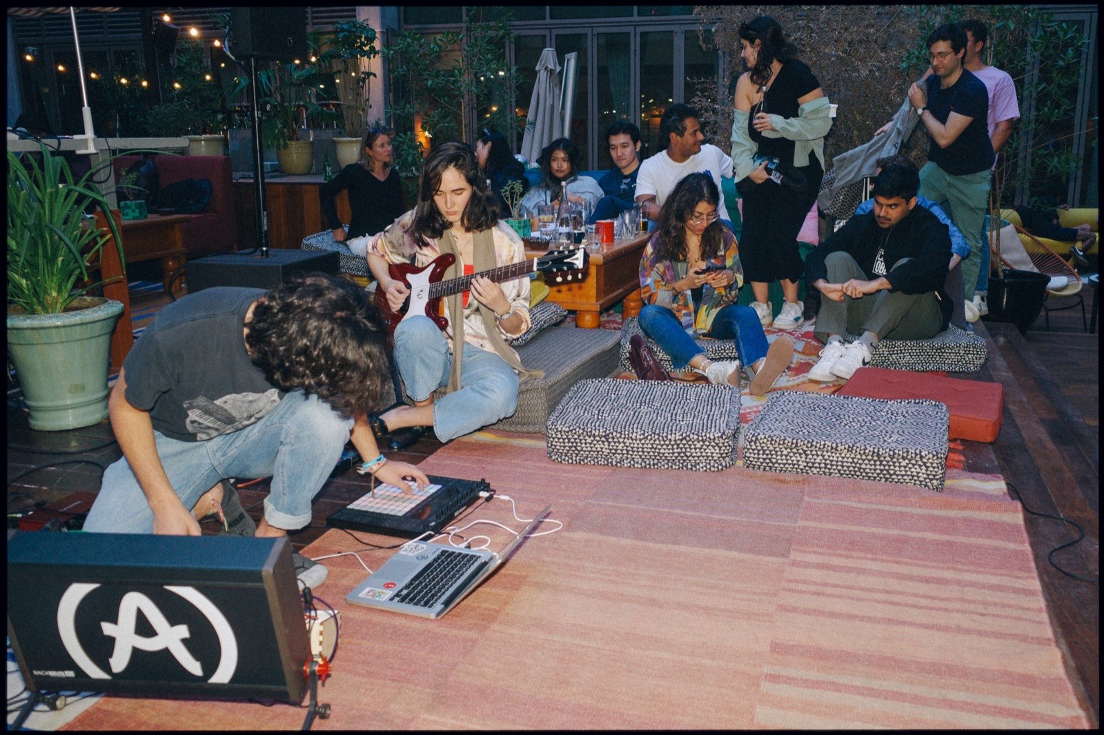 People sitting on rugs and cushions, listening on a terrace.