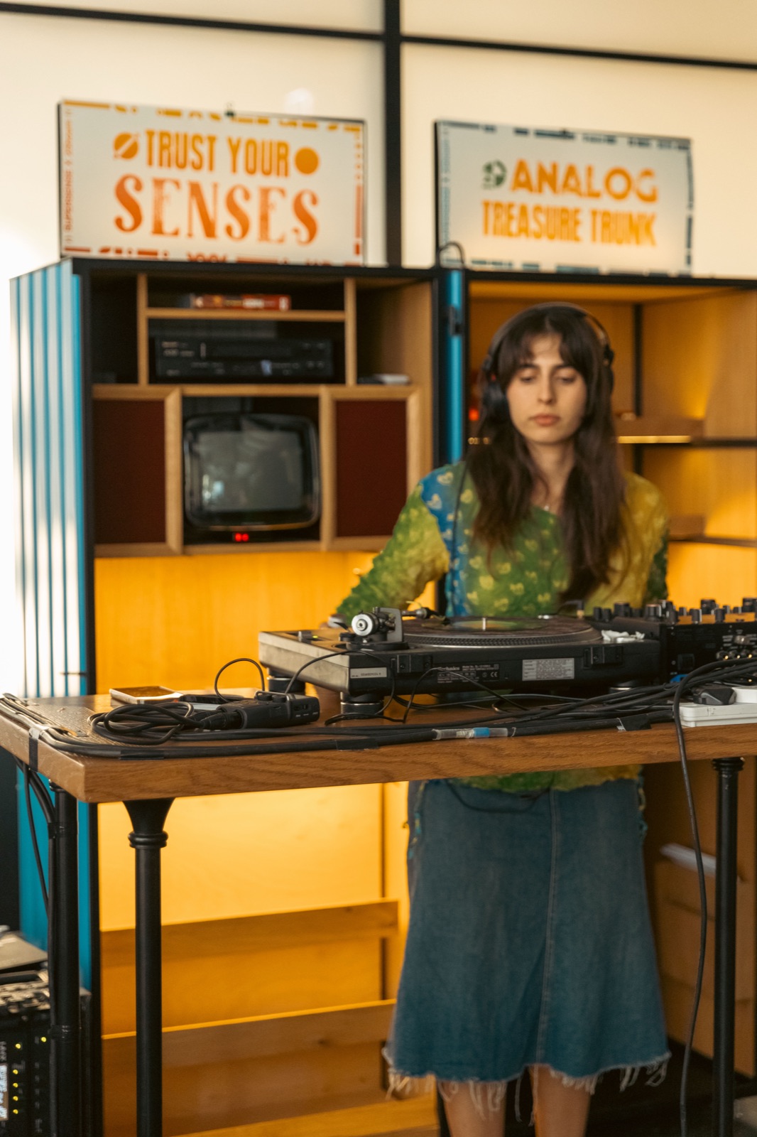 A DJ in a green top playing records in front of Trust Your Senses and Analog Treasure Trunk signs.