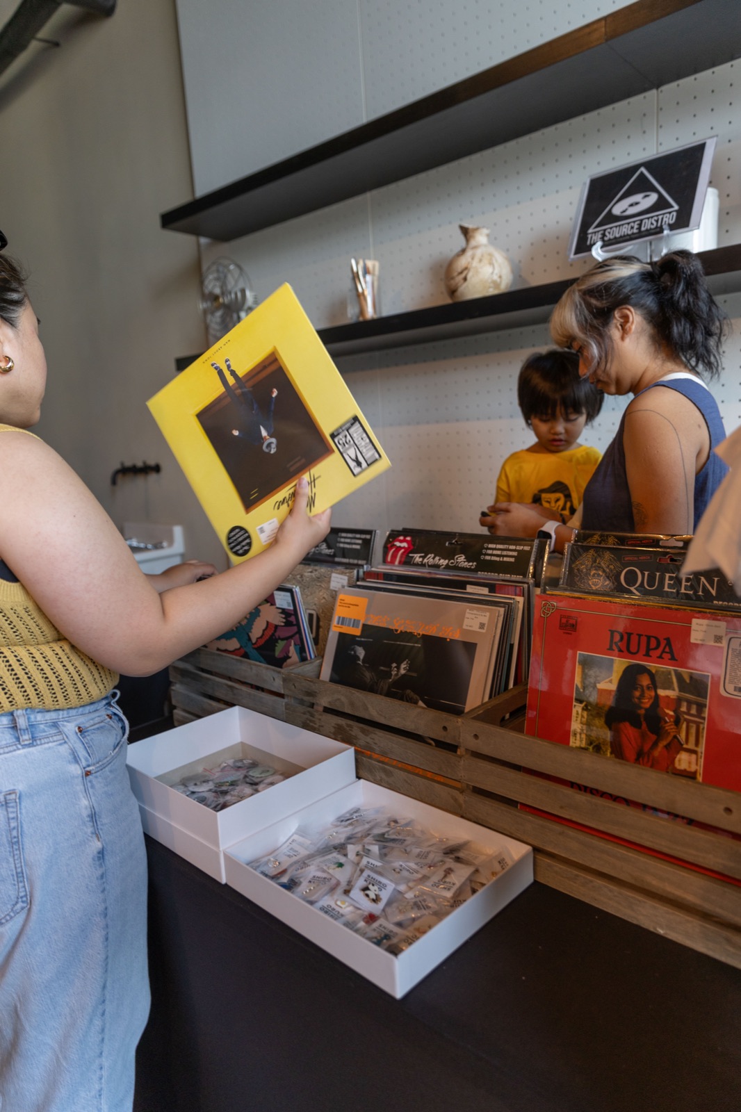 People browsing a record shelf, one holding up a yellow record sleeve.