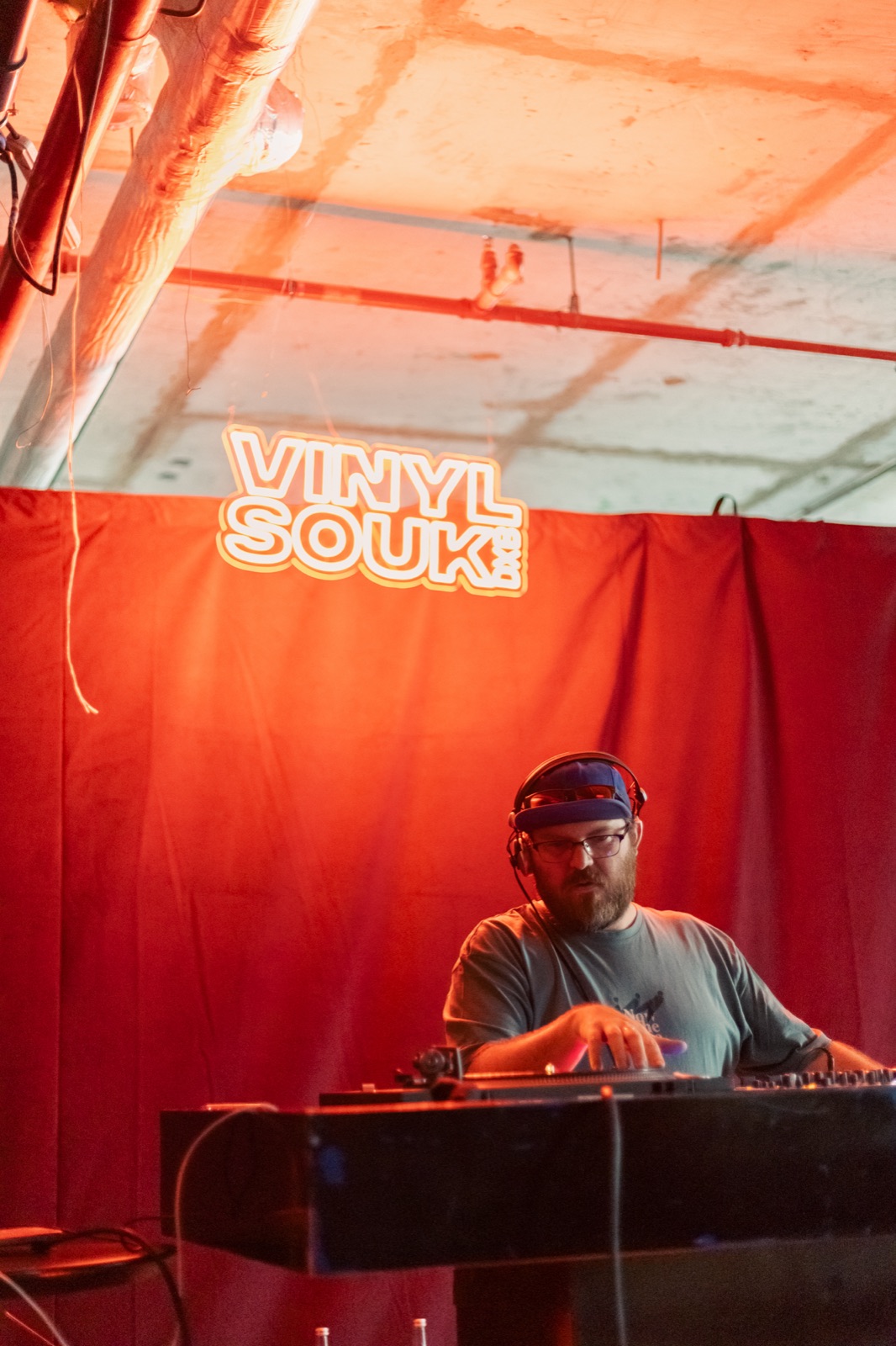 A DJ in a cap playing under a Vinyl Souk neon sign against a red curtain.