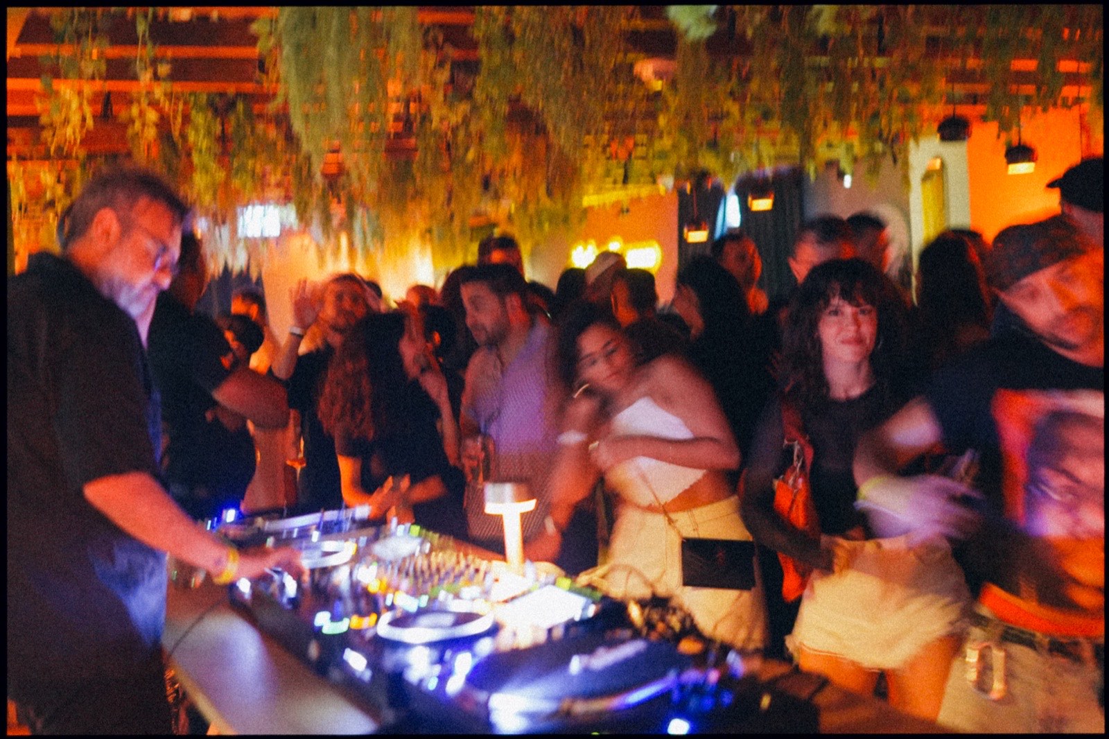A packed evening crowd around a DJ booth under hanging plants and warm lights.