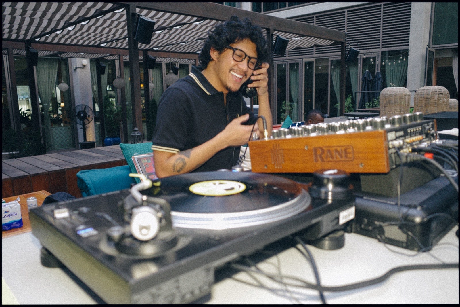 A DJ smiling at a turntable on a rooftop terrace, beside a rotary mixer.
