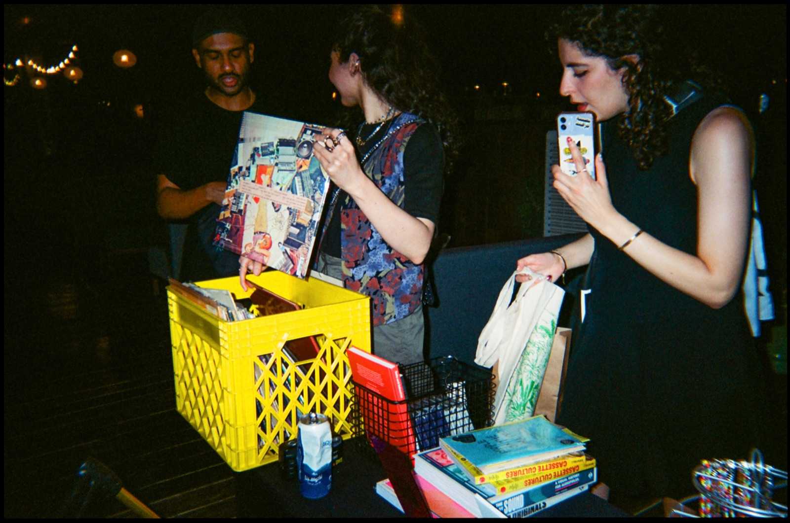 People browsing records from a yellow crate at night, one taking a photo with a phone.