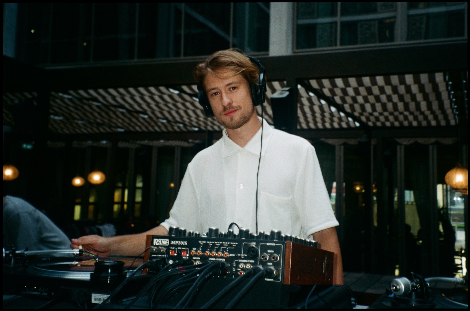 A DJ in headphones and a white shirt at the decks on a terrace at night.