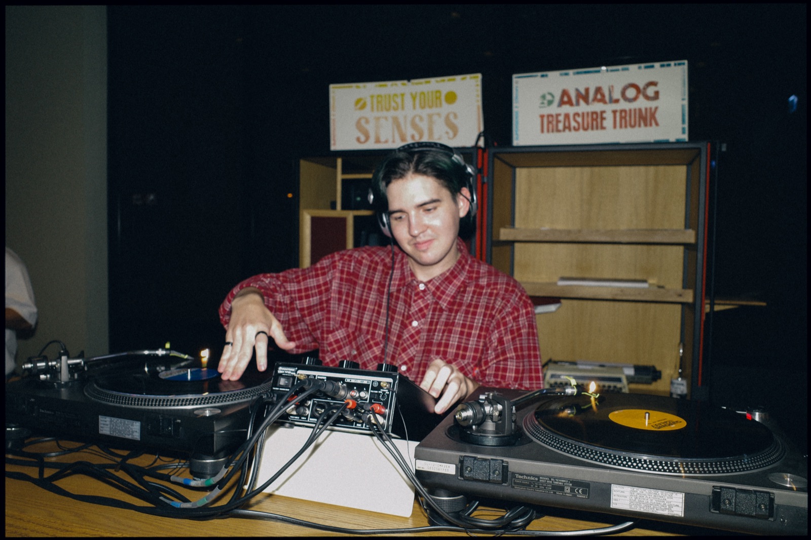 A young DJ in a checked shirt playing records under Trust Your Senses and Analog Treasure Trunk signs.