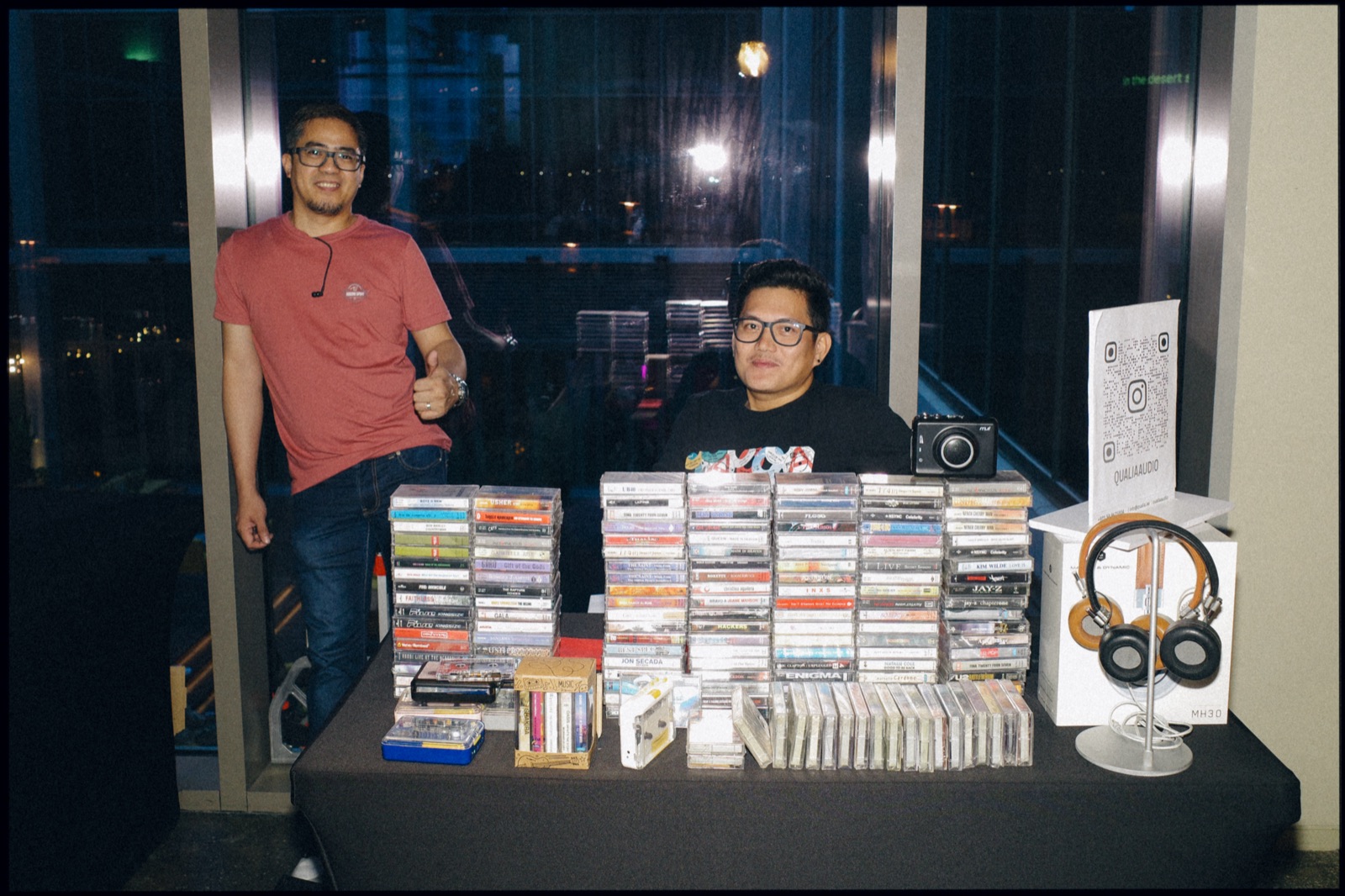 Two men behind a table of cassette tapes and headphones.
