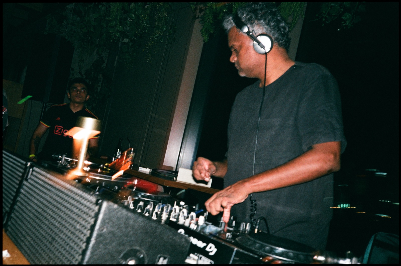 A DJ in headphones mixing in a dark booth lit by a small lamp.