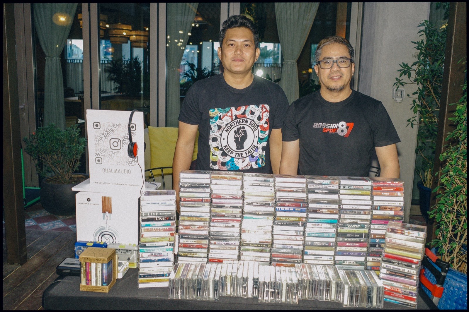 Two men behind a table stacked high with cassette tapes.