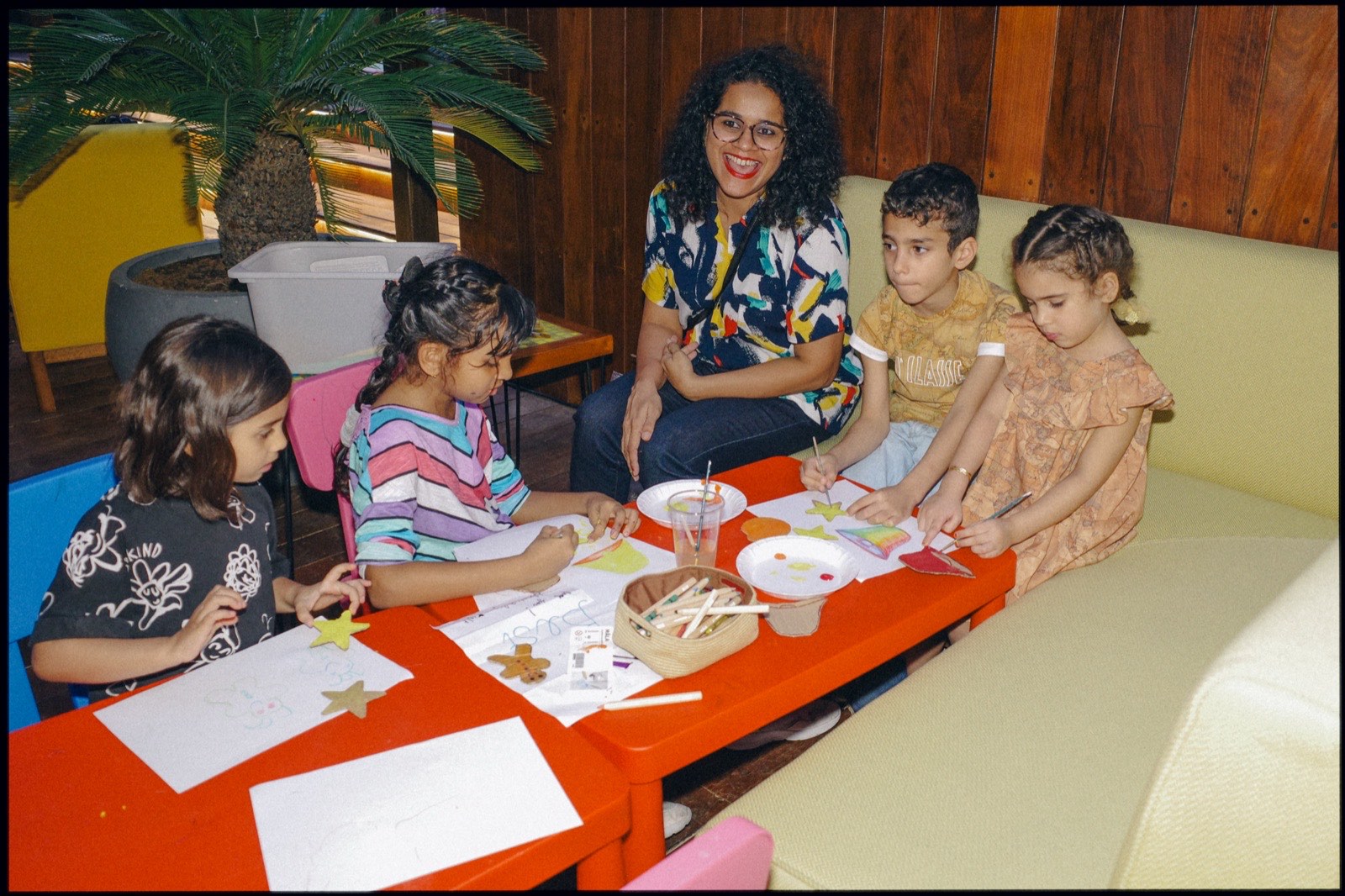 Children drawing at a red table, with an adult on the sofa.