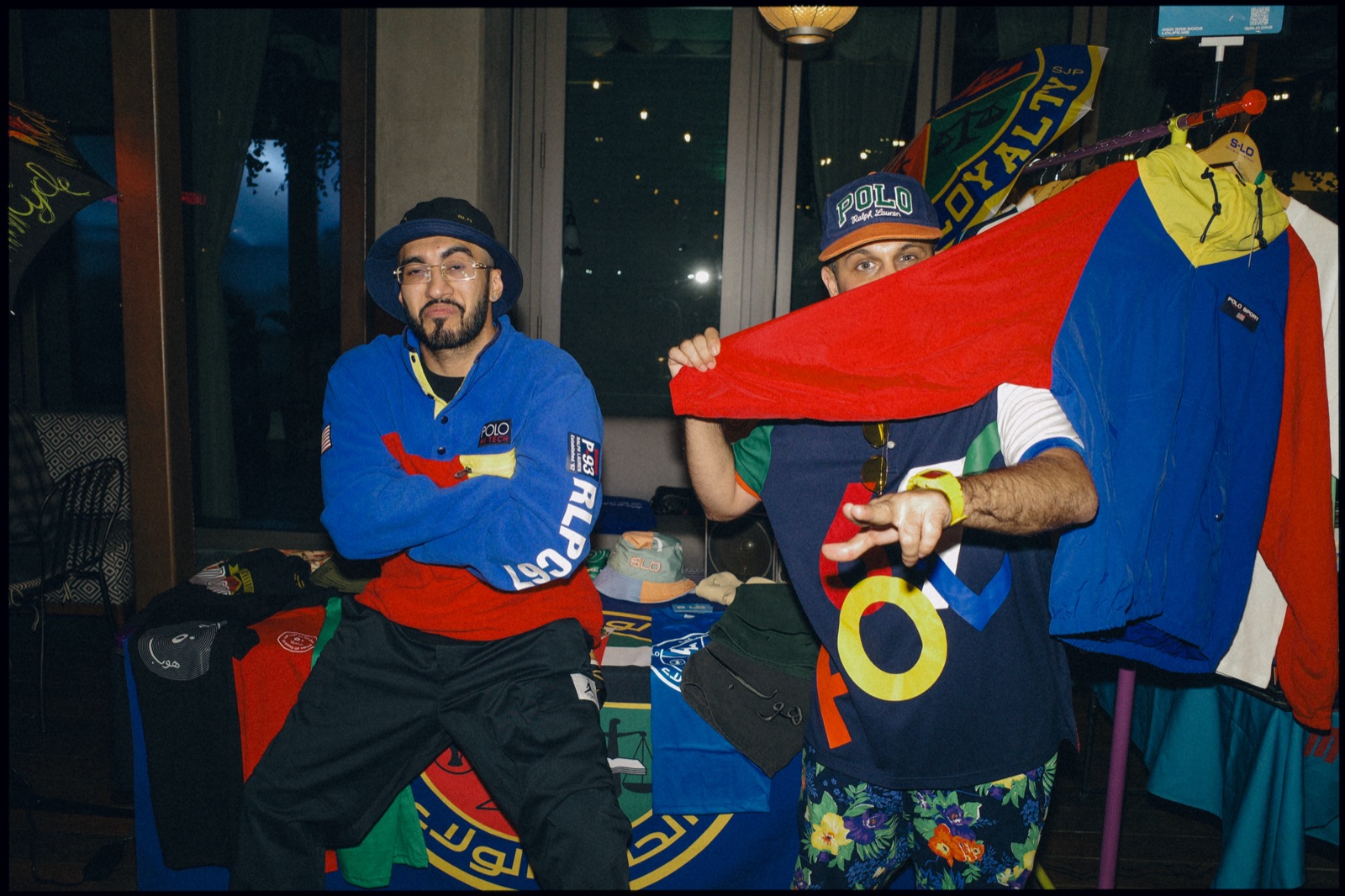 Two men posing in colourful retro sportswear beside a stall.