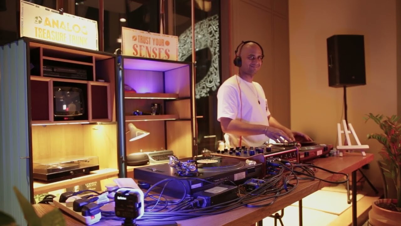 Watch on YouTube: a DJ in headphones playing records under Analog Treasure Trunk and Trust Your Senses signs, from Souk Sessions x 25h.